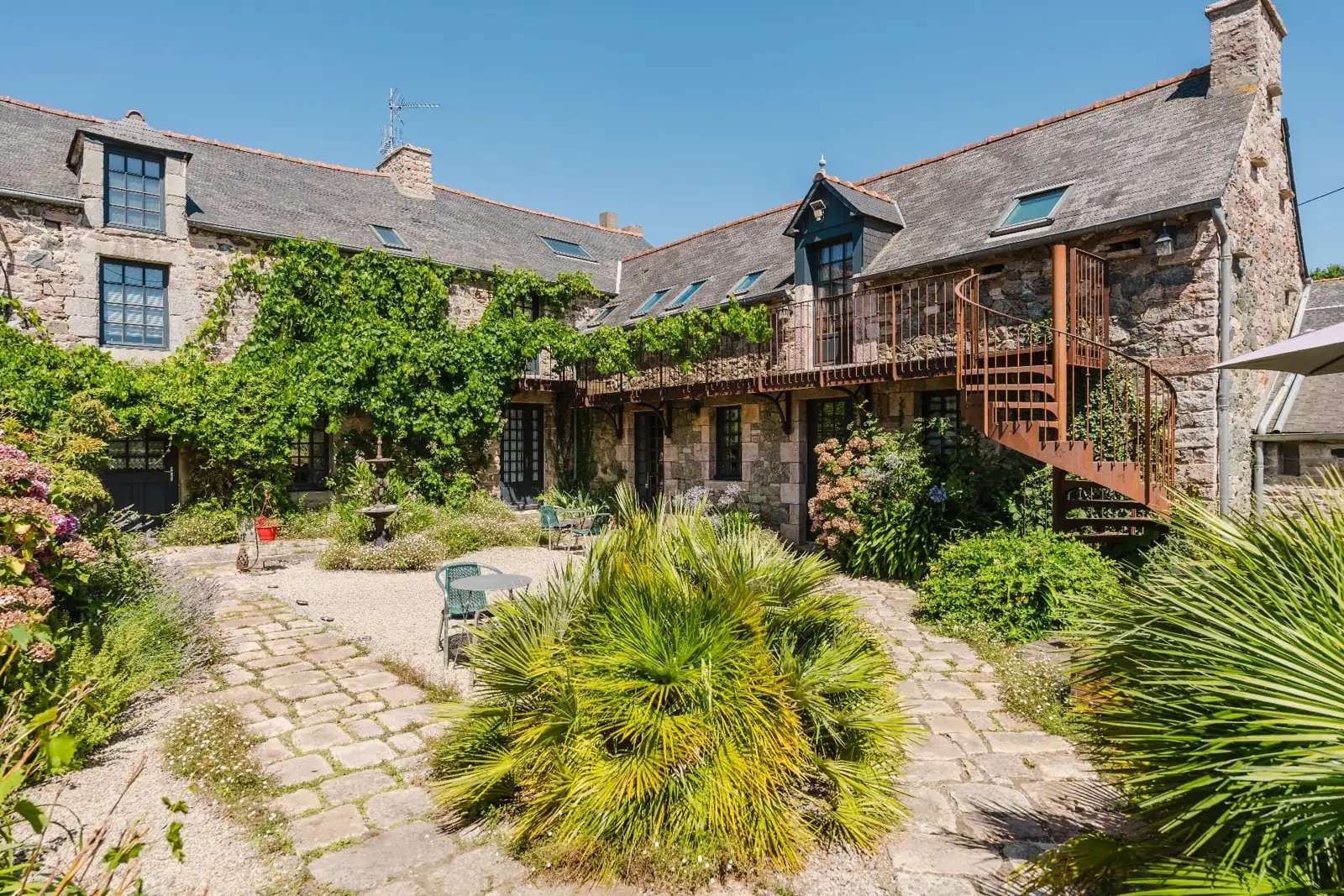 Cour pavée et fleurie du Relais Saint-Aubin, coursive en vigne et escalier en acier le long de la maison en pierre